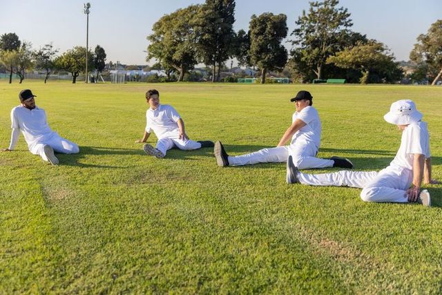 Cricket teammates stretching on sunlit outfield in white uniforms and caps before match