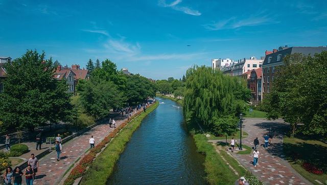 Sunlit urban canal promenade with willow trees, riverside walkways and walkers in summer
