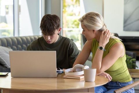 Woman helping teenage boy with laptop, tutoring and mentoring study session at home