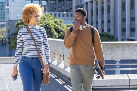 African american friends walking in city carrying skateboard and phone on sunny urban bridge
