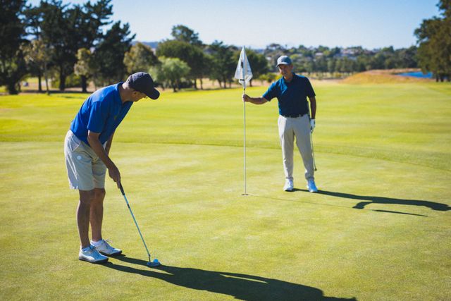 Two senior men are playing golf on a sunny day. One man is holding the flag while the other prepares to take a shot. This image can be used to promote healthy retirement lifestyles, outdoor activities for seniors, and golf as a hobby. It is ideal for advertisements, brochures, and articles related to sports, health, and leisure activities for older adults.