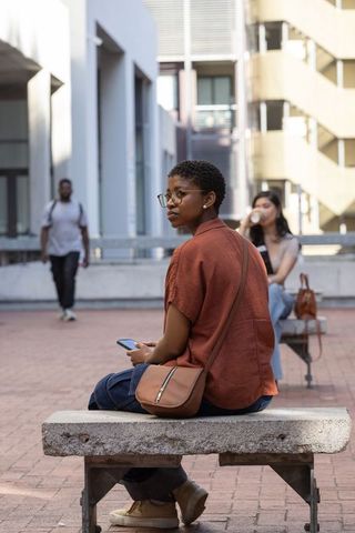 Woman sitting on bench holding smartphone in urban plaza, rust shirt and crossbody bag