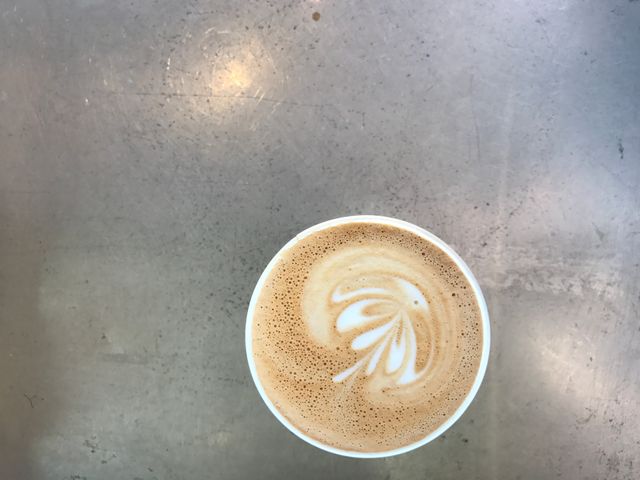 Overhead view of a cappuccino featuring intricate latte art on frothy milk, placed on a stainless steel table. Perfect casual concept for articles, coffee shop advertisements, or social media posts revolving around coffee culture, barista skills, or morning routines.
