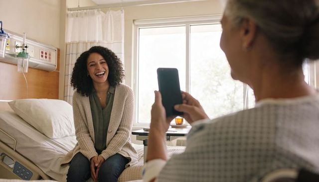 Friend laughing with patient while companion using smartphone in sunlit hospital room