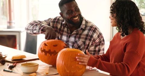 Couple carving jack-o-lanterns on sunlit table sharing autumn craft and cozy laughter