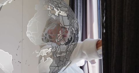 Senior woman reaching for curtain with globe overlay gazing toward window in soft light