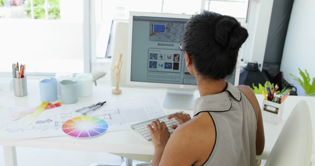 Female designer working at a modern desk with computer, engaged in creative project work. Includes color palette and design materials on desk. Ideal for illustrating concepts of work, creativity, design, technology in the workplace.