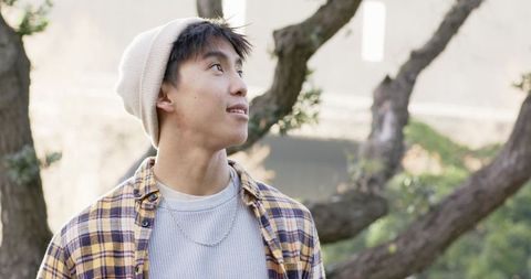 Young asian man wearing beanie and plaid shirt gazing upward among park trees, casual style