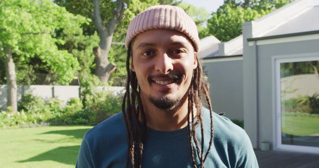 The image shows a young man with dreadlocks and a beanie smiling happily. He stands in a backyard garden with lush greenery and sunlight illuminating the scene. Perfect for use in lifestyle blogs, outdoor-themed advertisements, fashion campaigns for casual wear, or articles promoting positive vibes and healthy living.