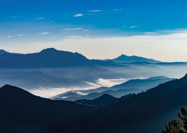 Depicting a serene morning scene with mist gently rolling over a mountain range under a clear bluish sky. Ideal for use in travel and tourism promotions, nature blogs, and websites focusing on outdoor adventures and scenic beauty. It can also be used as an inspirational background for websites or desktop wallpapers highlighting tranquility and natural beauty.