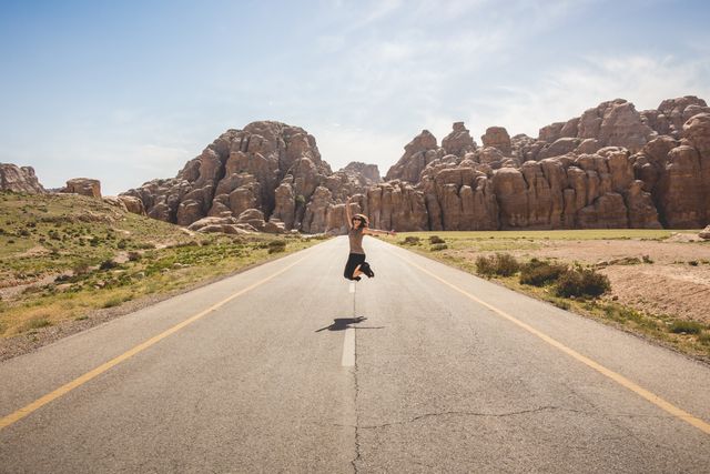 Woman jumping in excitement on an empty desert highway, surrounded by rocky formations under a sunny sky. Perfect for themes on travel, adventure, freedom, and solitude. Can be used in travel brochures, websites promoting road trips, inspirational blogs, or advertisements.
