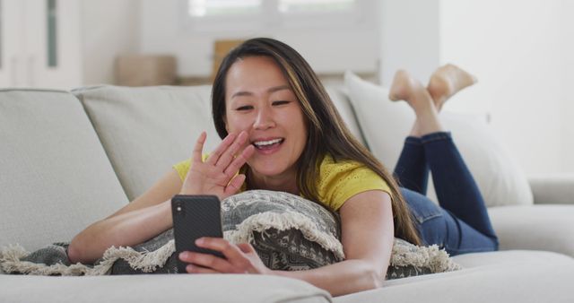 Woman lying on a comfortable sofa while video chatting on a smartphone. She appears relaxed and happy, enhancing the cozy and intimate atmosphere. This can be used for promoting communication applications, internet service providers, or home and lifestyle products.