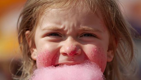 Young child biting pink cotton candy holding sticky sugar on sunlit cheeks at carnival