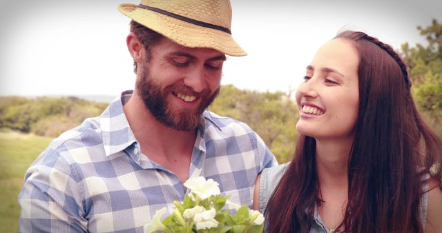 A young Caucasian couple shares a joyful moment outdoors, with the man holding a small bouquet of flowers, with copy space. Their smiles and casual attire suggest a relaxed, romantic setting, celebrating an intimate occasion or enjoying a date in nature.