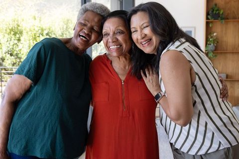 Laughing multiracial senior women hugging in sunlit modern living room
