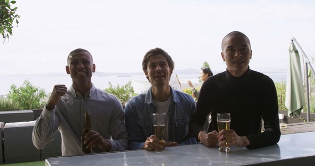 Three friends are enjoying their drinks at an outdoor bar with a scenic ocean view. They are smiling and appear to be having a good time. This can be used to illustrate concepts such as friendship, relaxation, celebration, and leisurely activities in a social setting.