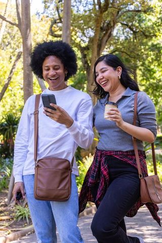 Young couple walking sunlit park laughing while checking smartphone and holding coffee