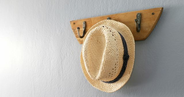 A straw hat hangs on a wooden rack against a white wall, with copy space. Its placement suggests a casual, homey atmosphere, often associated with warm weather or a countryside setting.