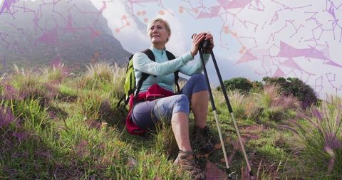 Senior woman resting and gazing on misty hillside with trekking poles and backpack