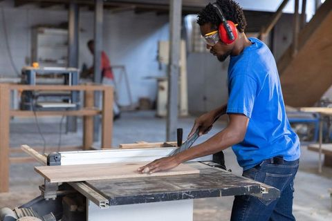 African American Carpenter Cutting Wood on Table Saw Wearing Safety Gear in Workshop