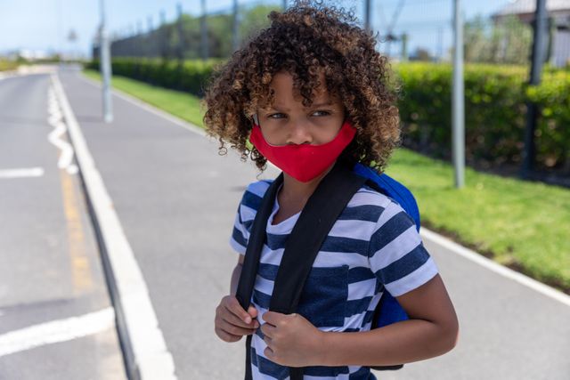 Young African American boy standing on the road wearing a face mask and backpack, emphasizing safety and health during the coronavirus pandemic. Ideal for use in educational materials, health and safety campaigns, and articles about school during COVID-19.