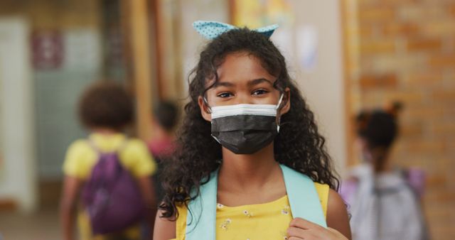 Portrait of biracial schoolgirl wearing face mask, standing in corridor looking at camera. children in primary school during coronavirus covid 19 pandemic.