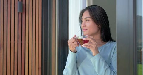 Mid-adult asian woman standing at sliding glass door holding teacup gazing peacefully