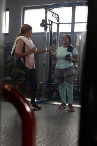 African american women chatting and smiling after workout in gym holding phone and bag