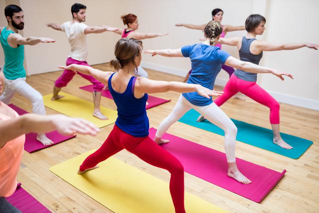 Group of people practicing yoga in a fitness studio, performing the Warrior Pose. They are standing on colorful yoga mats, stretching with their arms extended. The setting highlights a collaborative fitness environment. Perfect for promoting yoga studios, fitness classes, healthy lifestyle, group exercise programs, and wellness retreats.