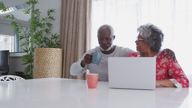 Senior African American couple spending quality time together at home, engaged in conversation while using a laptop. The man is holding and drinking from a mug, seated at a white table near a large window. Perfect for depicting themes of retirement, home life, quarantine, and technology usage among older adults.