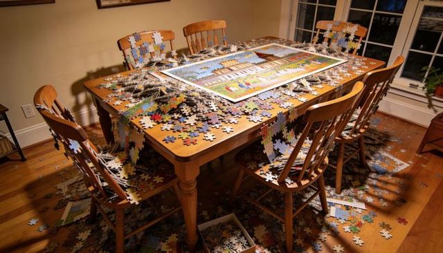 Overflowing jigsaw puzzle covering wooden dining table, spilling pieces on chairs and floor