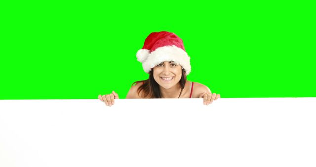 Woman in Santa hat smiling and holding blank banner against green background. Useful for holiday-themed promotions, Christmas advertising, and festive messages. Ideal for digital creations where viewers can easily place their own text or images on the blank banner.