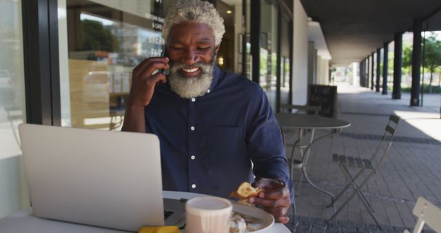 Senior man with grey hair enjoying breakfast while using a mobile phone and laptop at an outdoor cafe. Ideal for depicting senior lifestyle, communication, technology, and business themes. Suitable for advertising products targeting mature individuals, showcasing casual business interactions, or promoting cafes and dining experiences.