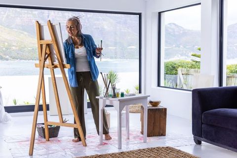 Senior woman painting at easel in bright waterfront studio wearing headphones