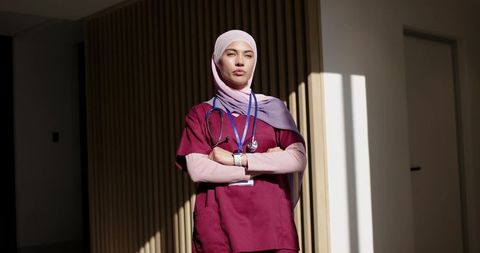 Confident muslim nurse standing in sunlit clinic corridor wearing scrubs and stethoscope