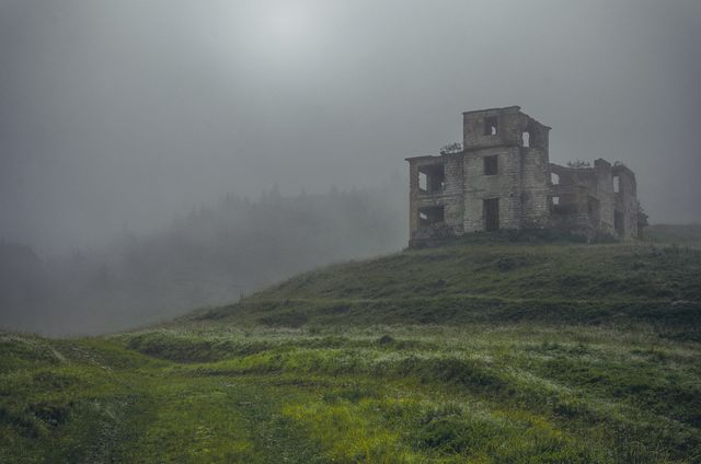 This image features an abandoned building standing alone on a foggy hill in a misty countryside. The eerie atmosphere and desolate landscape create a spooky and mysterious vibe, ideal for use in horror stories, spooky settings, and environmental pieces highlighting the beauty of forgotten places. The overgrown nature and forest in the background add depth and mystique, making it suitable for themes of abandonment and nature reclaiming human structures.