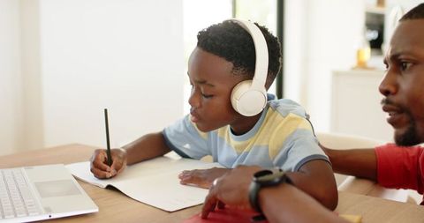 African american father supporting son studying at table while child wearing headphones