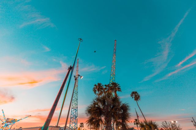 Amusement park rides rise against a vibrant sunset sky adorned with palm trees, creating a picturesque and energetic scene. Ideal for promoting vacation destinations, leisure activities, amusement parks, travel brochures, and summer events.