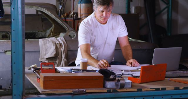 Caucasian man reviews plans in a workshop setting. He's focused on his project, surrounded by tools and a laptop, illustrating hands-on work.