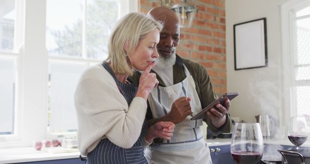 Biracial senior couple wearing aprons using digital tablet while cooking in the kitchen at home. retirement senior couple lifestyle living concept