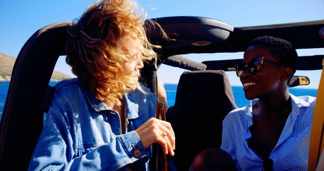 Two friends enjoying a road trip in a convertible car on a sunny day by the beach. They are chatting happily, one holding a bottle, both with carefree expressions. Ideal for lifestyle, travel, and adventure advertising, youth-focused campaigns, and summer vacation stories.