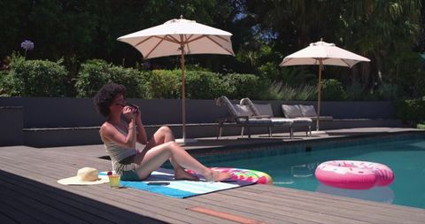 African american woman relaxing poolside under umbrellas adjusting sunglasses with sunhat