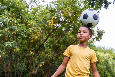African boy balancing soccer ball on head in park wearing yellow shirt showing focus