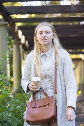 Young woman walking under pergola holding takeaway coffee and brown leather handbag