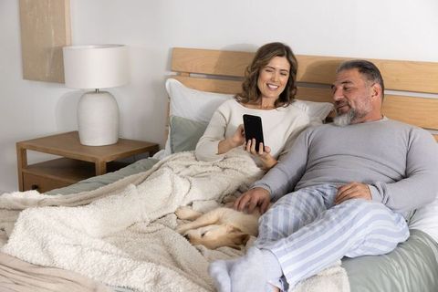 Middle-aged couple relaxing in bed sharing smartphone and petting dog under cozy blanket