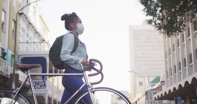This image shows a woman standing in an urban environment while holding a bicycle and wearing a face mask. Ideal for illustrating themes related to cycling, urban commuting, pandemic safety measures, and healthy lifestyles. Suitable for use in articles, advertisements, and websites focused on urban life, health precautions, and eco-friendly transportation.
