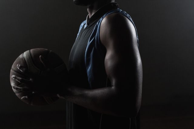 This image captures a basketball player holding a basketball against a dark background, emphasizing the athlete's muscular arm and determination. Ideal for use in sports-related content, fitness promotions, motivational posters, and athletic training materials.