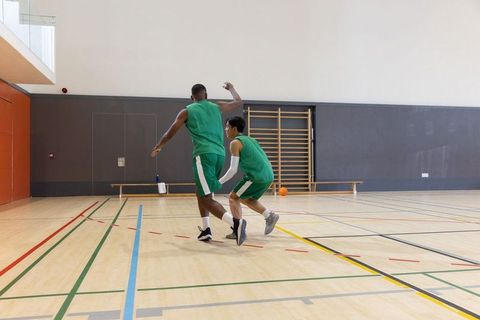 Diverse teammates practicing defensive footwork while dribbling basketball on gym court
