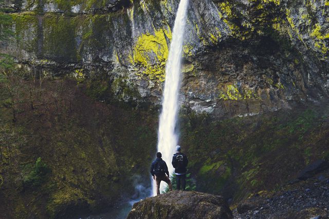 This stunning image captures two hikers standing in front of a massive waterfall cascading down rocky cliffs, illustrating a sense of adventure and the beauty of the natural world. Perfect for travel ads, outdoor adventure promotions, or nature-inspired content, emphasizing scenic exploration and outdoor activities.