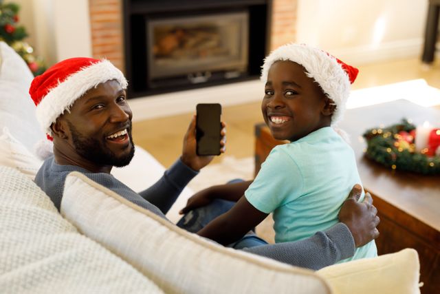 Rearview of an african-american man and his son wearing santa hats sitting on the couch looking behind them with smiles on their faces. the man is holding a phone up.